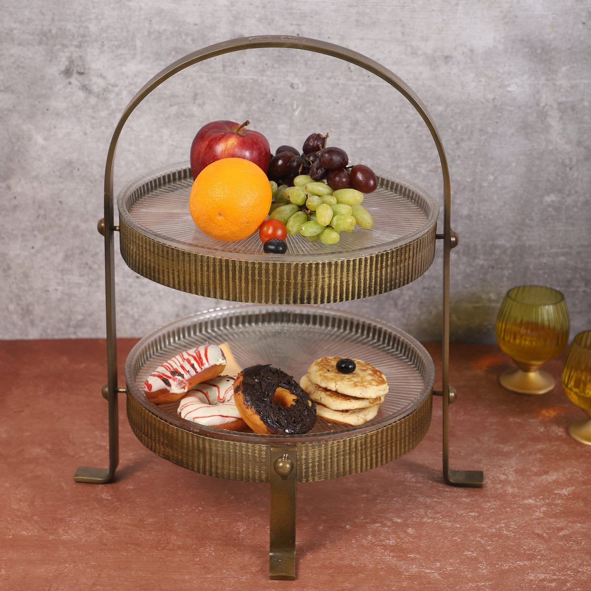 TTwo-tiered vintage-style metal organizer with ribbed gold finish, holding skincare bottles, a blue perfume bottle, a yellow candleholder, and small potted plants, placed on a marble surface.