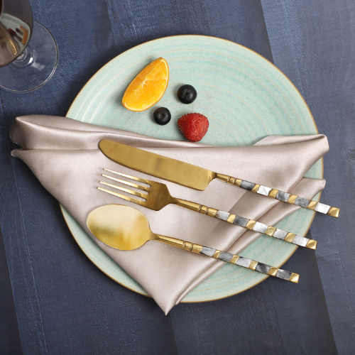  A decorative table setting featuring gold flatware with silver handles on a beige napkin, placed on a light blue ceramic plate, accompanied by fruit arranged to resemble a face with blueberries, a strawberry, and an orange slice