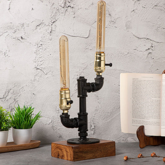 Industrial-style table lamp with a pipe-inspired design, dual brass-tone bulb holders, and a wooden base, displayed alongside greenery and an open book on a concrete-textured background